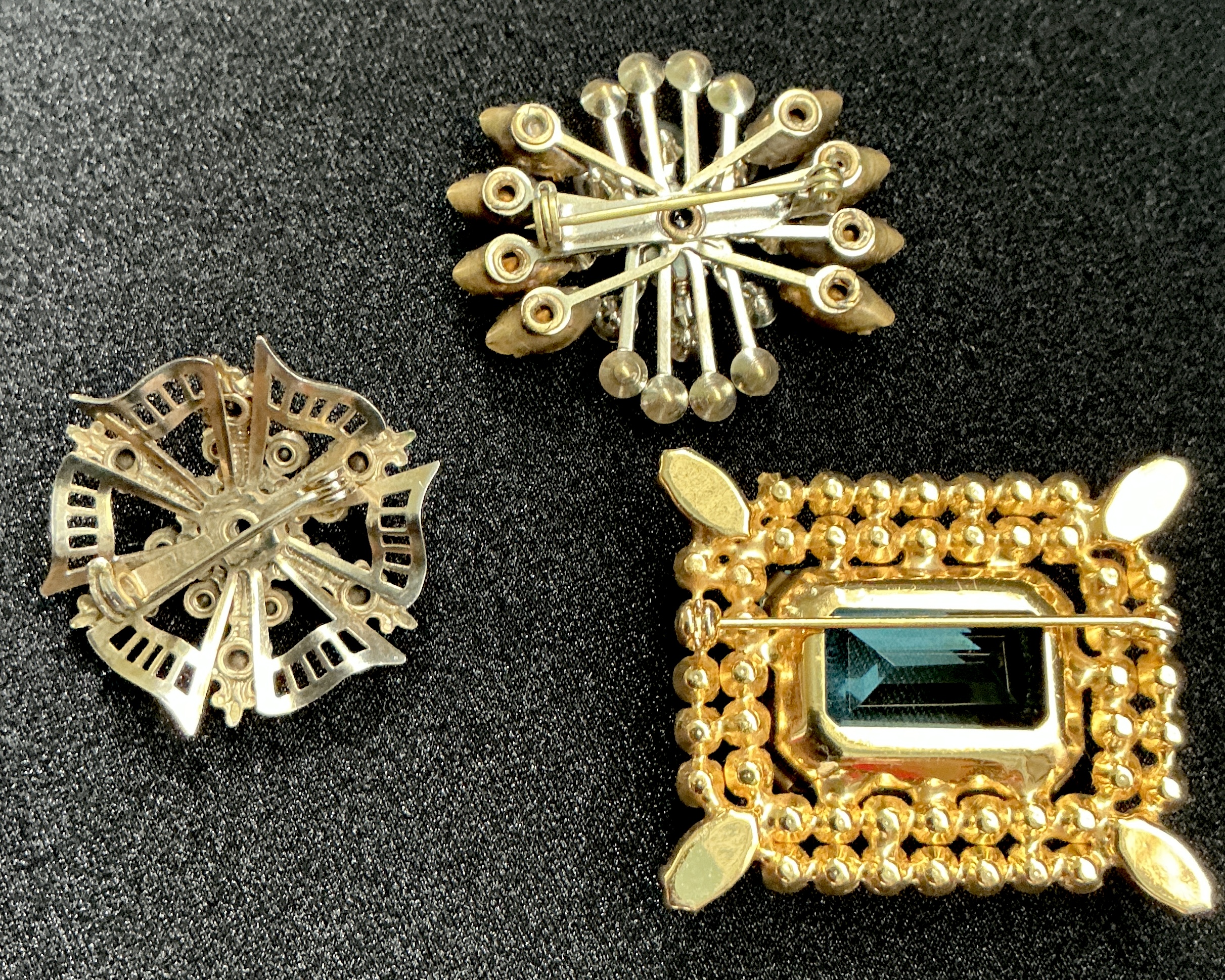 Backs of three vintage brooches: a starburst, a snowflake, and a rectangular emerald-style brooch.